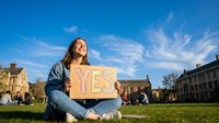 Young woman of mixed ethnicity holding yes sign on grass in sunny public square, expressing positive decision and agreement for lifestyle, education, or community concepts.