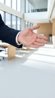 Caucasian male professional in dark business suit extending open hand for handshake or greeting in modern office environment with natural light and contemporary workspace.