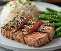 Grilled sesame-crusted tuna steak sliced and served medium rare with steamed rice and green beans on white plate, garnished with fresh herbs and Asian seasonings.