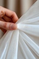 Female hand with nude manicure gently holding delicate white tulle fabric, creating soft folds and texture. Close-up view shows fine mesh detail and ethereal draping.