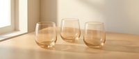 Three elegant amber-tinted stemless wine glasses arranged on wooden surface with warm natural sunlight creating soft shadows in minimalist interior setting.