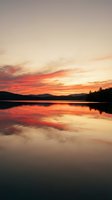 Serene lake at sunset with vibrant orange and red sky reflecting on calm water surface, surrounded by distant mountains and evergreen forest silhouettes.