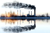 Industrial power plant with smoking chimneys reflecting in calm water at sunrise, dark smoke clouds against winter sky creating mirror effect on lake surface.