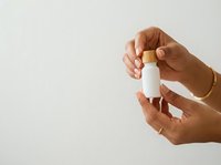 Person with medium-dark skin tone holding white cosmetic bottle against neutral background for skincare, beauty product, or wellness branding and packaging mockup purposes.