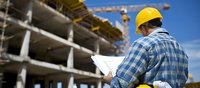 Male construction supervisor in yellow hard hat reviewing blueprints at building site, wearing plaid shirt against concrete structure and blue sky background. For construction industry.