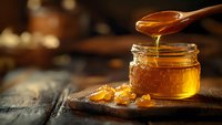 Golden honey dripping from wooden spoon into glass jar on rustic wooden table with warm bokeh background for natural food and organic products.