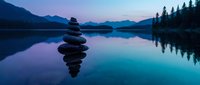 Zen stone stack balanced on calm lake water at twilight with mountain silhouettes and pine trees reflecting in serene wilderness landscape.