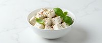 Fresh mozzarella cheese cubes with basil leaves and black pepper in white bowl on marble surface for culinary presentations and food photography.