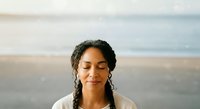 Young woman with dark braided hair and South Asian ethnicity, eyes closed in peaceful expression, wearing white clothing against soft beach and ocean backdrop, conveying calm and mindfulness.