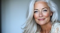 Elegant mature woman with silver hair and natural makeup smiling confidently against blurred background. Genuine expression showcases aging gracefully and feminine beauty.