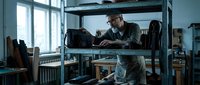 Male craftsman working at workbench in workshop, organizing tools and materials on metal shelving unit near window with natural light for carpentry, woodworking, or manufacturing projects.