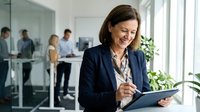Smiling mature businesswoman using digital tablet in modern office with colleagues working in background during daytime meeting.