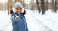 Happy young African American girl in winter coat and knit hat pointing at camera in snowy park during sunny day for seasonal lifestyle content.
