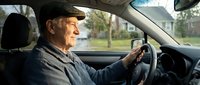 Senior Caucasian man driving car with confident smile, elderly driver behind wheel enjoying road trip, mature male motorist in vehicle interior.