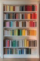 Colorful books arranged by spectrum on white built-in bookshelf, creating rainbow pattern across six shelves. Classical home library interior with hardcover volumes.