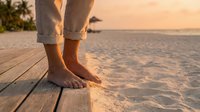 Barefoot person standing on wooden dock at sunset, feet touching sandy beach with ocean waves in background, warm golden hour lighting, relaxation and vacation concept.