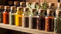 Colorful spices and herbs in glass jars with cork lids arranged on wooden shelf for cooking, seasoning and culinary preparation in kitchen pantry.