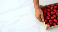 Farmer's hand holding wooden crate filled with fresh red strawberries on white marble surface, harvest and organic produce concept for food marketing.