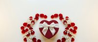 Heart-shaped gift box with white ribbon surrounded by red rose petals on light background for Valentine's Day romantic celebration and love concept.