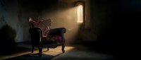 Ornate vintage armchair positioned in a dimly lit room with dramatic golden light streaming through a tall narrow window, creating atmospheric shadows and highlighting the upholstered furniture in an abandoned interior setting.