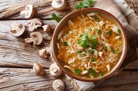 Homemade chicken noodle soup with mushrooms and fresh herbs served in rustic ceramic bowl on wooden table, comfort food perfect for restaurant menu or cooking blog.