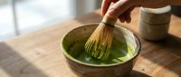 Matcha tea preparation with whisk in ceramic bowl on wooden surface, hand whisking green powder into hot water for traditional Japanese beverage making.