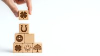 Hand placing wooden cube with four-leaf clover symbol on pyramid of blocks featuring lucky symbols including horseshoe and lotus on white background.