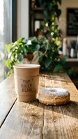 Steaming coffee in kraft paper cup with glazed donut on wooden table in bright cafe interior with green plants and natural light, perfect for food and beverage marketing.