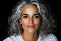 Confident mature woman with natural gray wavy hair and radiant smile against dark background showcases authentic aging and self-acceptance in modern portrait.