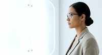 Young woman of East Asian descent wearing eyeglasses and beige blazer, standing in profile beside modern glass partition wall in bright office environment, professional business setting.