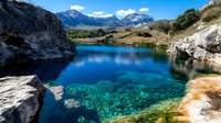 Pristine alpine lake with crystal clear turquoise water reflecting snow-capped mountains, surrounded by rocky terrain and green hills under blue sky.