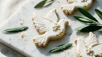Elegant dove-shaped sugar cookies with white royal icing and gold pearl accents on marble surface with olive branches for wedding celebrations.
