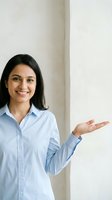 Smiling Hispanic businesswoman in blue shirt presenting with open palm gesture in modern office space for corporate communication and marketing materials.