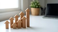 Wooden house blocks arranged in ascending growth pattern on white desk with laptop and plant in modern office workspace for real estate investment concept.
