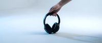 Hand holding black over-ear wireless headphones against a clean light blue-gray background, showcasing modern audio technology.