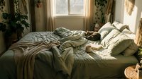 Cozy bedroom with unmade bed, natural sunlight streaming through window, potted plants, and warm neutral tones for home decor and lifestyle content.