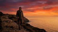 Middle-aged male hiker with walking stick stands on rocky coastal cliff overlooking golden sunset ocean. Adventure and travel concept for outdoor lifestyle marketing.