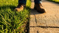 Close-up of brown leather boots on concrete pathway with green grass growing along edges, outdoor walking scene for lifestyle and nature concepts.