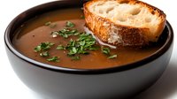 Rich brown soup garnished with fresh herbs and toasted bread slice in a dark ceramic bowl on white background.