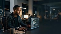 Focused male software developer with dark skin tone working late at computer in modern office, typing code on monitor, professional tech environment for business and technology concepts.