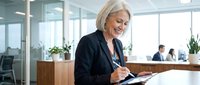 Mature blonde businesswoman writing notes in modern office environment with natural lighting and professional atmosphere.