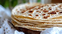 Stack of homemade flour tortillas with golden brown spots on white cloth, perfect for Mexican cuisine preparation.