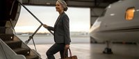 Professional blonde businesswoman in dark suit boarding private jet with luggage at airport terminal during golden hour for corporate travel and luxury lifestyle.