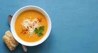 Creamy orange soup in white bowl with parsley garnish and bread roll on blue wooden table background for healthy comfort food recipes.