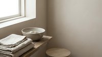 Minimalist bathroom corner with stacked folded linen towels, ceramic bowl and wooden stool by window with natural light for spa and home decor.