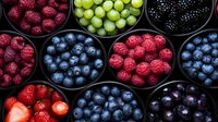 Fresh assorted berries in black bowls including raspberries, blueberries, blackberries, strawberries and green grapes arranged in circular pattern on dark background.