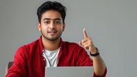 Young South Asian male professional in red cardigan points upward while working on laptop, expressing positive business idea or solution against gray background.