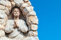 Religious sculpture in stone arch shows praying figure wearing white robe against bright blue sky, depicting spiritual devotion and architectural detail.