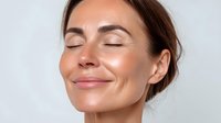 Caucasian woman with closed eyes and serene smile showing glowing, hydrated skin against light gray background, demonstrating skincare results.