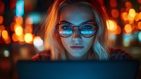 Young blonde woman in glasses illuminated by blue laptop screen light against dark background with orange bokeh lights, creating cyberpunk atmosphere.